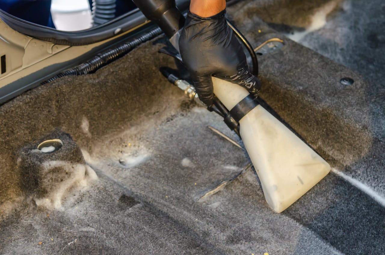 why-choose-us A hand in a glove using a vacuum to clean a car interior carpet, removing dirt effectively.